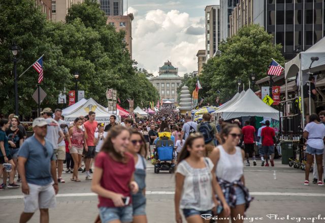 Crowded Raleigh festival with people walking among tents and vendors, American flags, and trees lining the sidewalks under a cloudy sky.