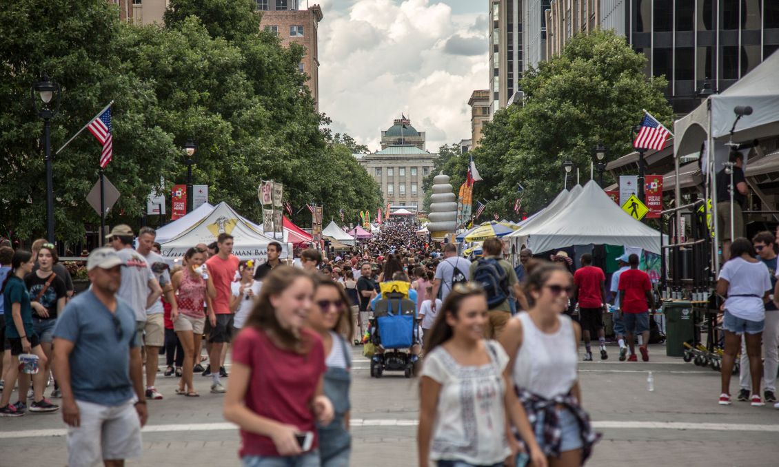 Crowded Raleigh festival with people walking among tents and vendors, American flags, and trees lining the sidewalks under a cloudy sky.