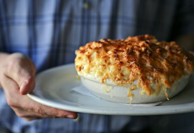 Person holding a white plate with a hot, golden-brown baked macaroni and cheese in a white casserole dish at Poole's Diner.