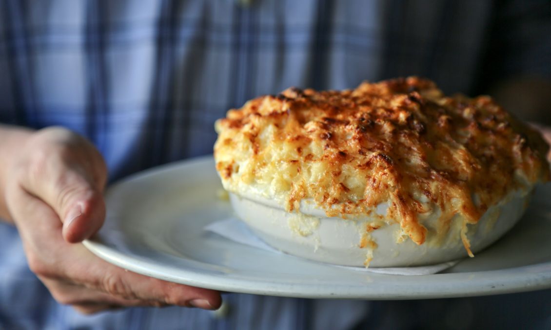 Person holding a white plate with a hot, golden-brown baked macaroni and cheese in a white casserole dish at Poole's Diner.