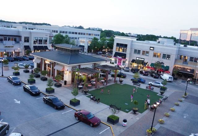 Aerial view of an outdoor shopping center in Raleigh with parked cars, a central pavilion, seating, and people walking and playing on a green lawn.