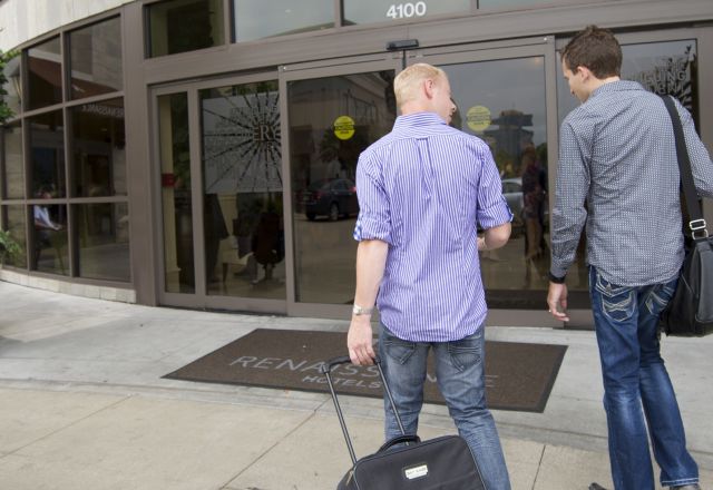 Two people walking toward the entrance of a modern hotel in downtown Raleigh with planters and city storefronts lining the sidewalk.