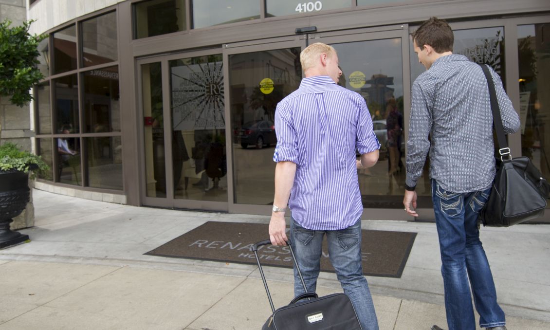 Two people walking toward the entrance of a modern hotel in downtown Raleigh with planters and city storefronts lining the sidewalk.