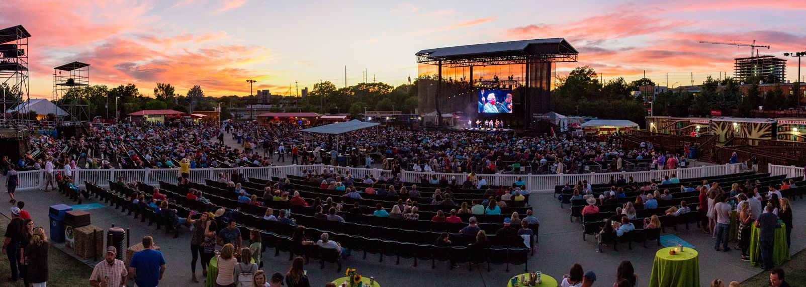 Outdoor music venue at sunset in Raleigh, NC, with a large crowd watching performers on an illuminated stage and a colorful sky backdrop.