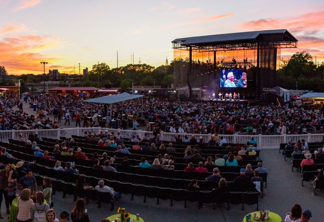 Outdoor music venue at sunset in Raleigh, NC, with a large crowd watching performers on an illuminated stage and a colorful sky backdrop.