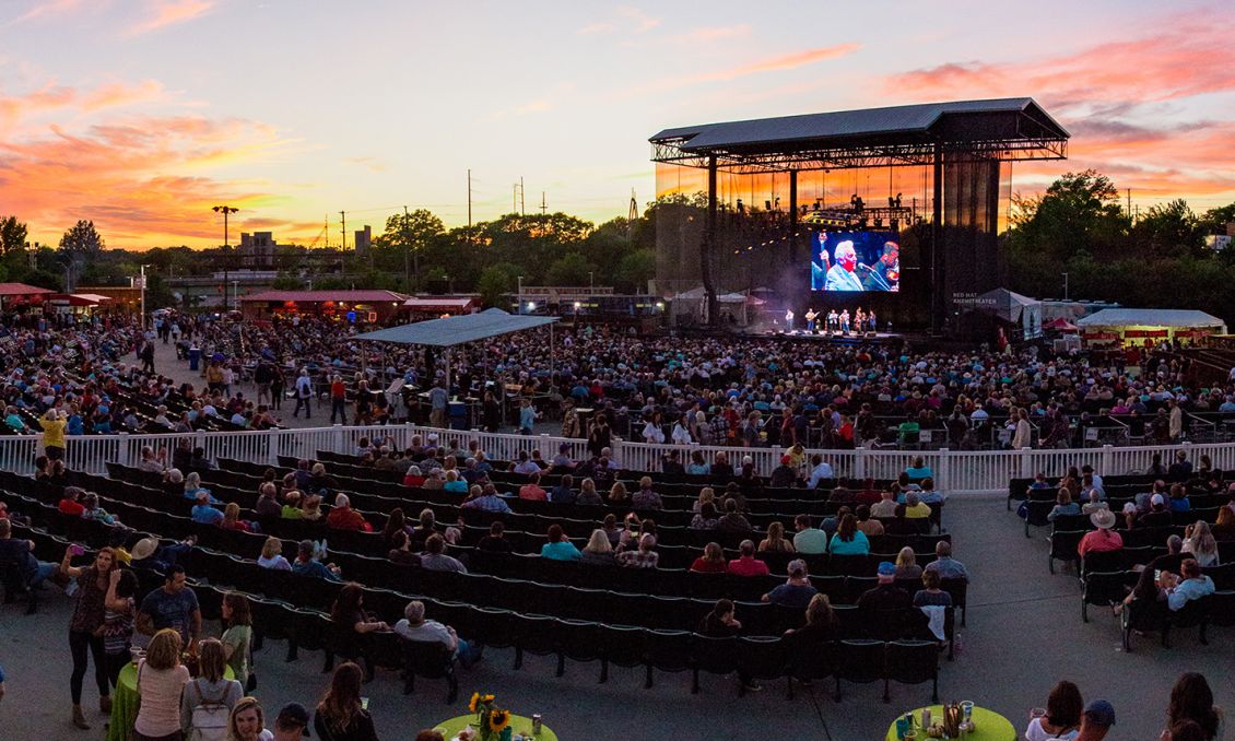 Outdoor music venue at sunset in Raleigh, NC, with a large crowd watching performers on an illuminated stage and a colorful sky backdrop.