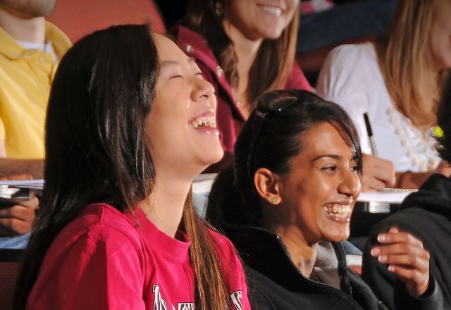 A group of college students seated in a lecture hall with red chairs, smiling while listening during class on campus setting today.