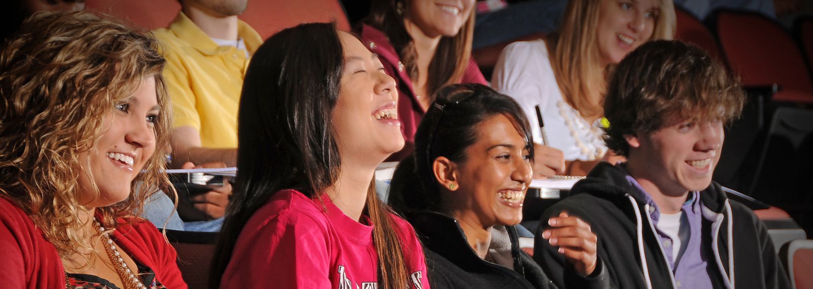 A group of college students seated in a lecture hall with red chairs, smiling while listening during class on campus setting today.