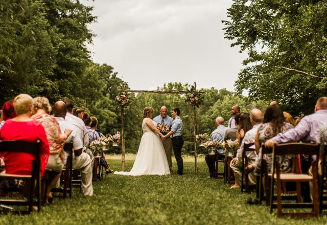 Outdoor wedding ceremony in Raleigh, NC, with the couple at the altar, guests seated on chairs, green lawn, trees, and rustic backdrop scene.