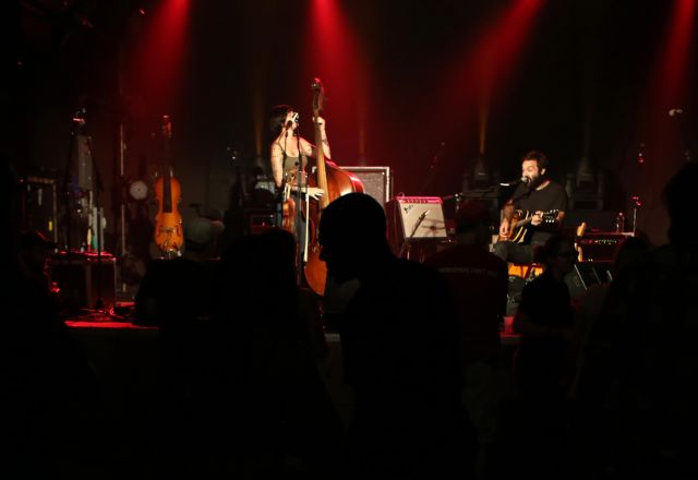 Live music performance with a double bass player and guitarist on stage under red spotlights, audience silhouettes in foreground.