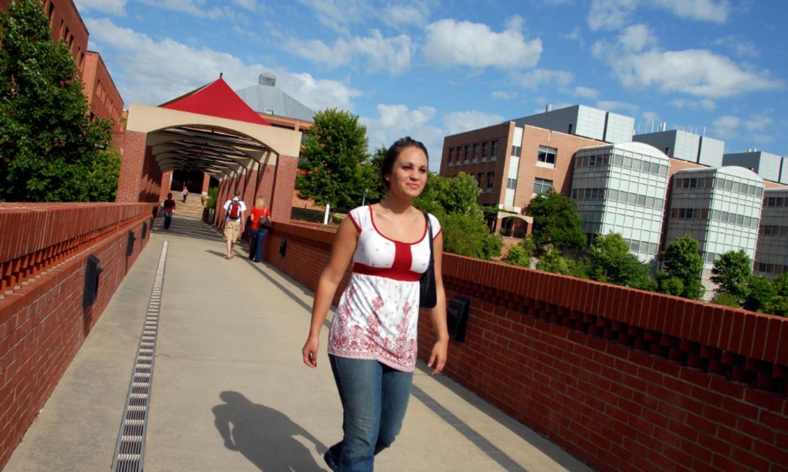 Student walking on North Carolina University Centennial Campus brick pathway with buildings, trees, blue sky, and a modern bridge.
