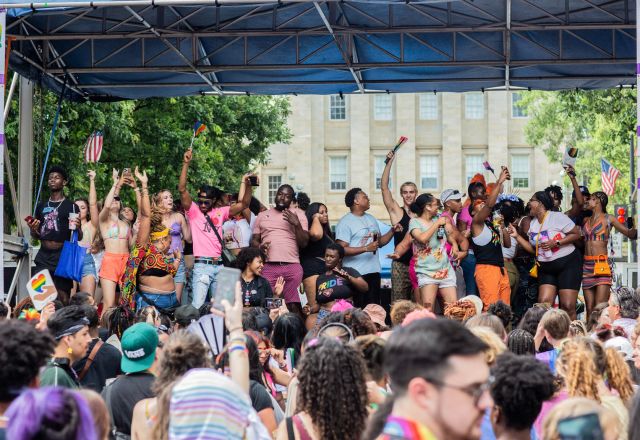 People gathered at Out Raleigh Pride stage with crowd dancing colorful flags and banners downtown festival atmosphere, live scene.